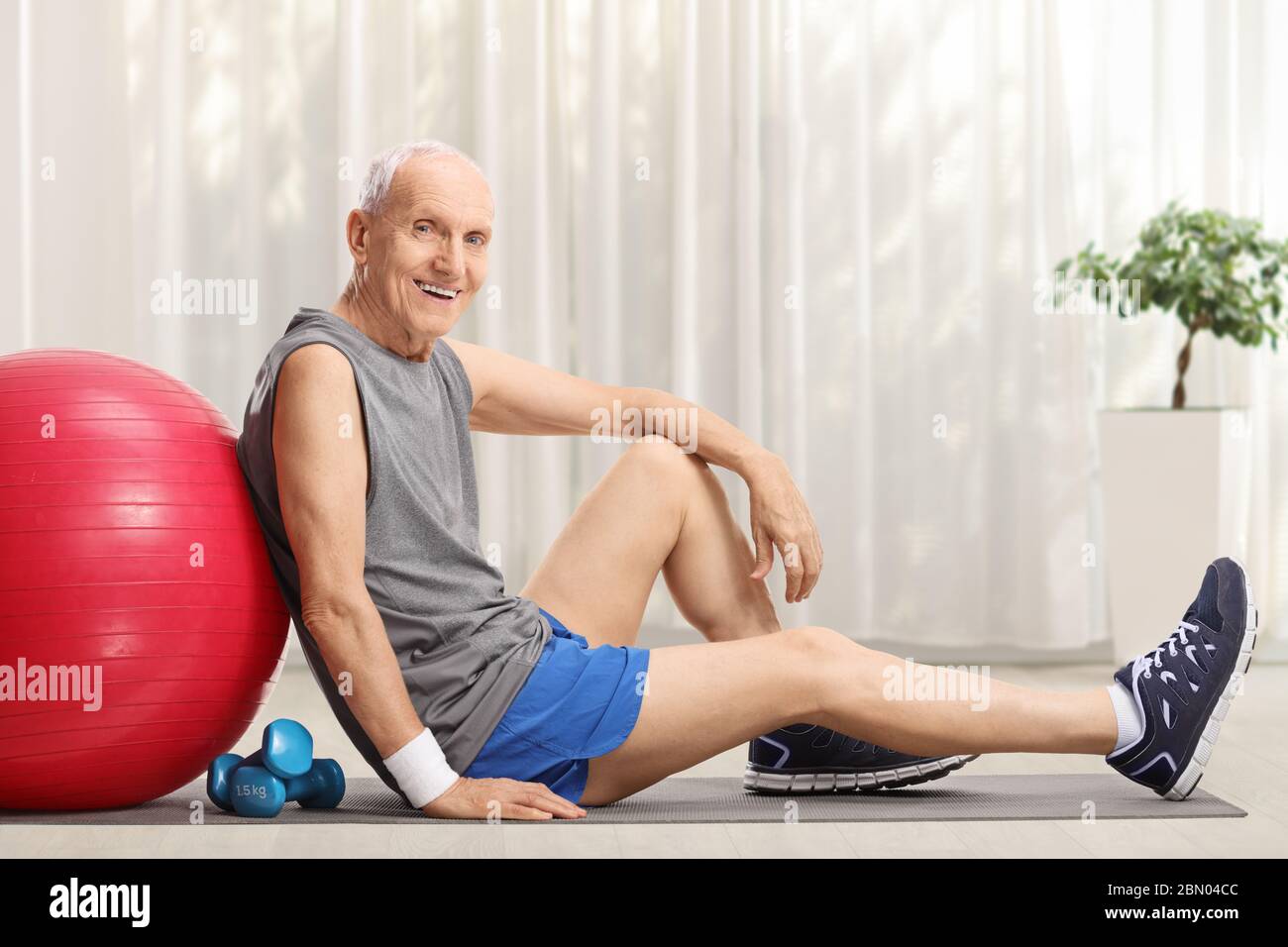 Älterer Mann mit Fitnessball und Hanteln sitzt zu Hause auf einer Trainingsmatte Stockfoto
