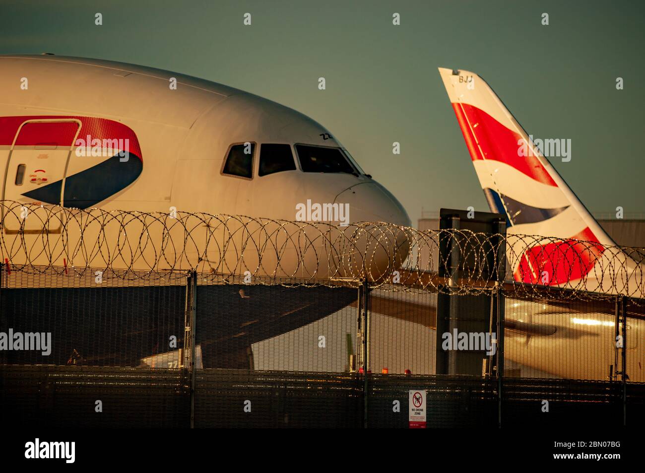 Die Nase eines Flugzeugs von British Airways neben dem Schwanz eines anderen hinter einem Stacheldrahtzaun und ohne Drohnenschild bei Sonnenuntergang. Stockfoto