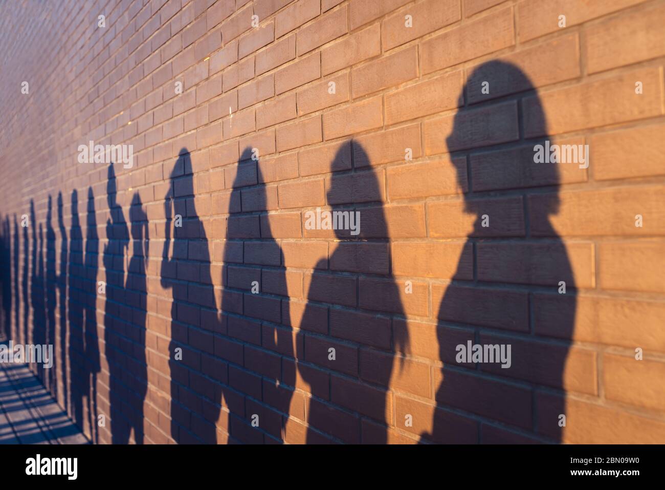 Die Menschen drängen Schatten aufgereiht gegen eine rote Ziegelmauer. Sie stehen in einer Schlange für Veränderungen im Leben. Soziale Distanz, Covid und Immigration Themenkonzept Stockfoto