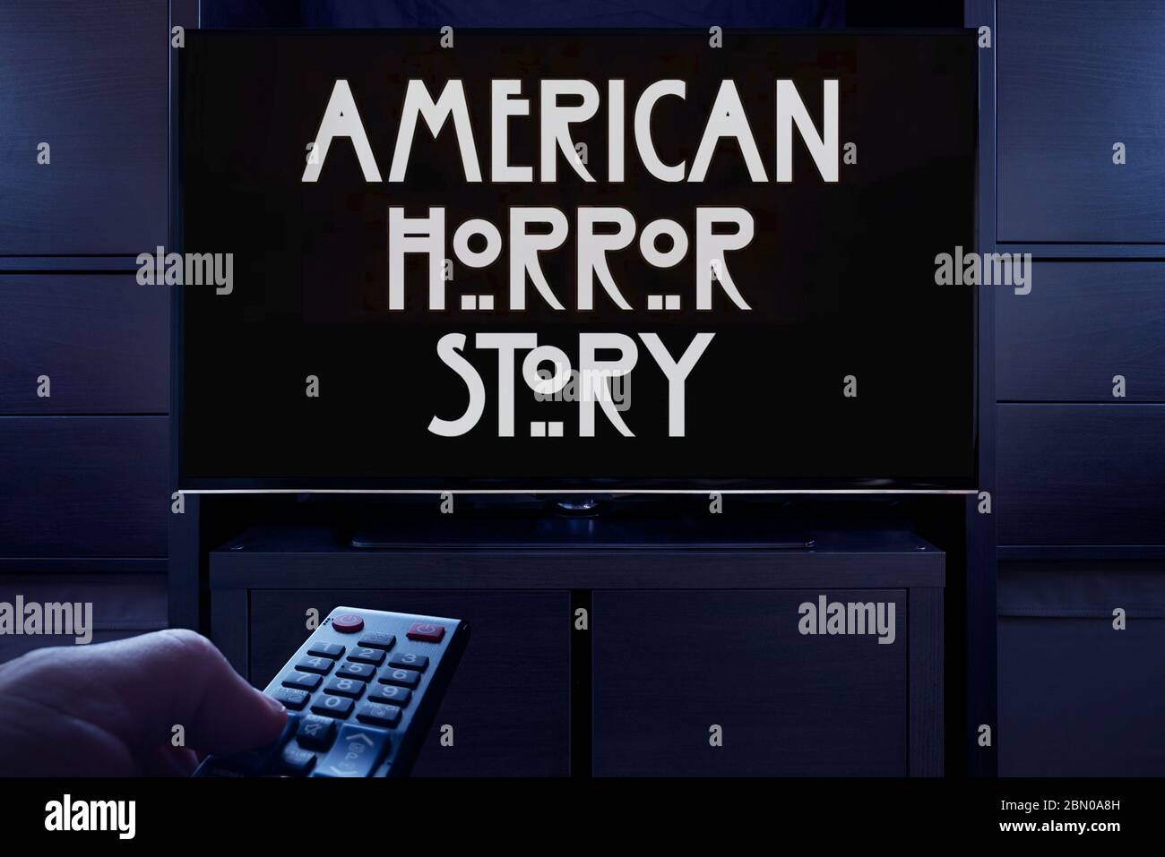 Ein Mann zeigt eine Fernbedienung auf den Fernseher, der den Haupttitelbildschirm der American Horror Story anzeigt (nur redaktionelle Verwendung). Stockfoto