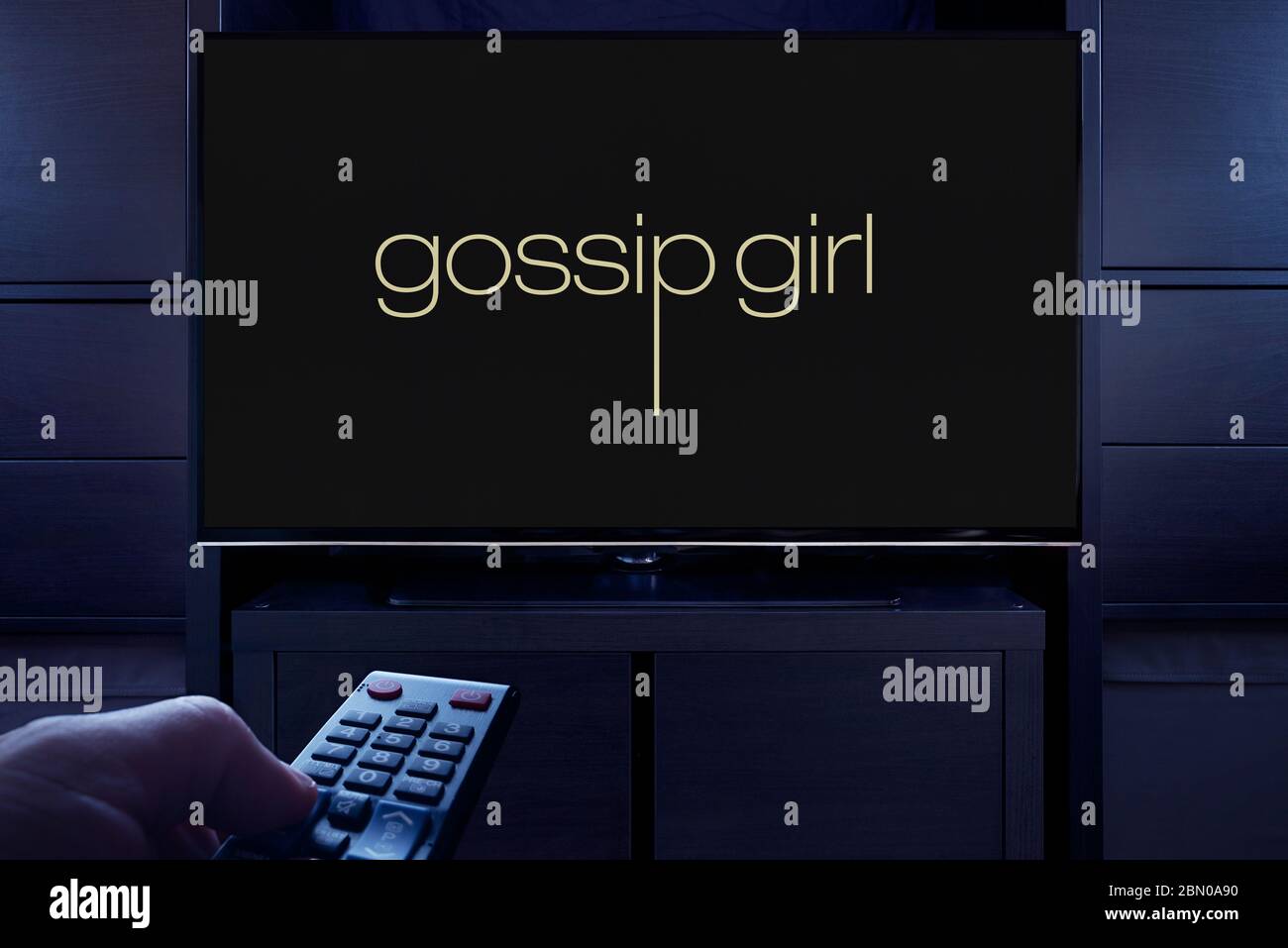 Ein Mann zeigt eine Fernbedienung auf den Fernseher, der den Hauptbildschirm von Gossip Girl anzeigt (nur für redaktionelle Zwecke). Stockfoto