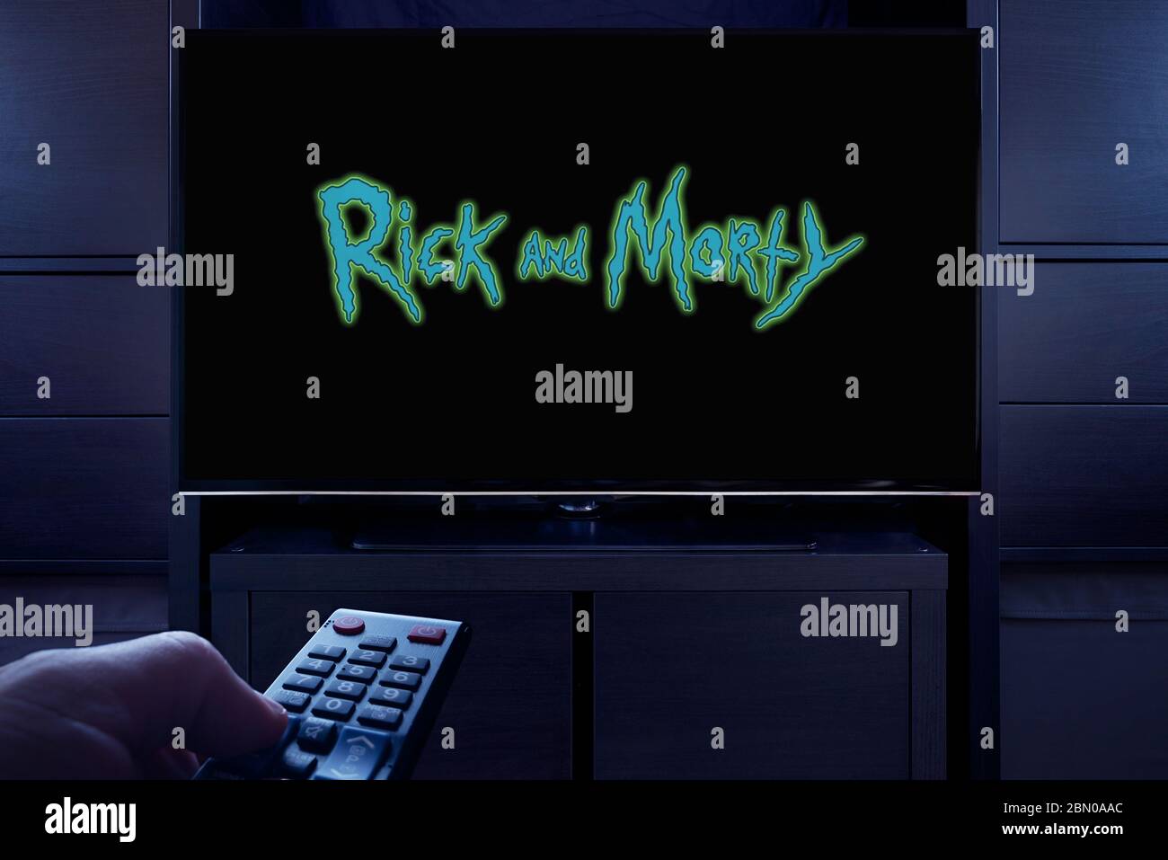 Ein Mann zeigt eine Fernbedienung auf den Fernseher, der den Hauptbildschirm von Rick and Morty anzeigt (nur redaktionelle Verwendung). Stockfoto