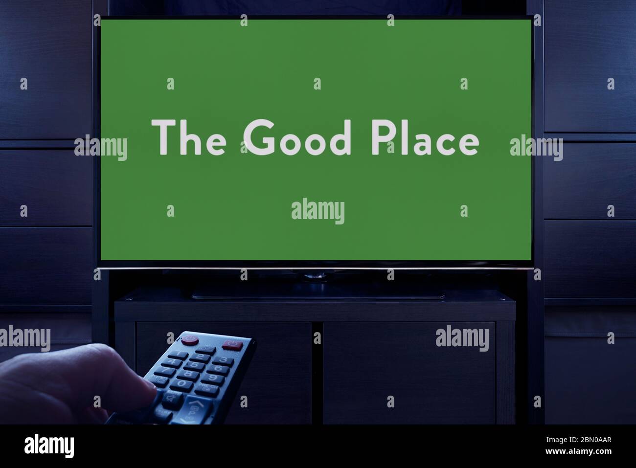Ein Mann zeigt eine Fernbedienung auf den Fernseher, der den Hauptbildschirm des guten Ortes anzeigt (nur redaktionelle Verwendung). Stockfoto