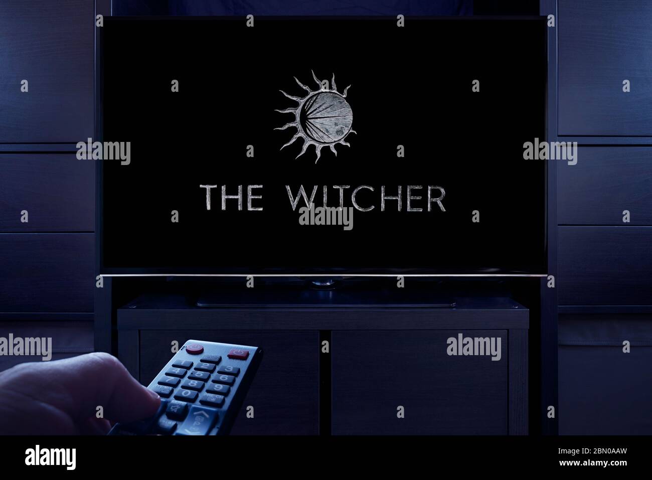 Ein Mann zeigt eine Fernbedienung auf den Fernseher, der den Hauptbildschirm des The Witcher-Titels anzeigt (nur redaktionelle Verwendung). Stockfoto