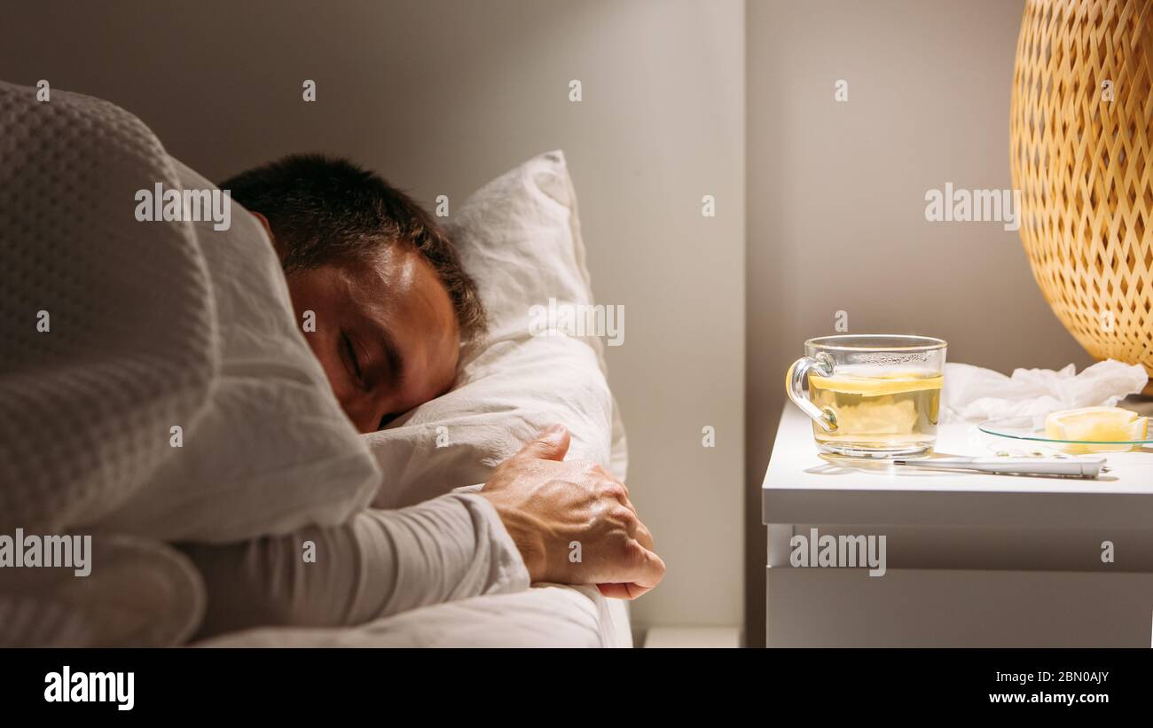 Kranker Mann schläft mit einer Decke bedeckt, die im Bett mit hohem Fieber liegt und im Wohnzimmer ruht. Er ist erschöpft und leidet an Grippe mit einer Tasse Tee Wi Stockfoto