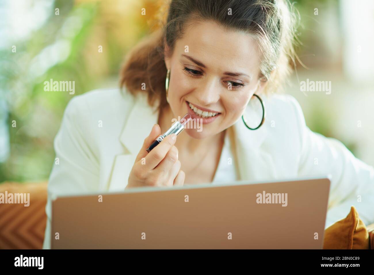 Happy modern Mittelalter Frau in weißer Bluse und Jacke mit Laptop Anwendung Lippenstift während auf der Couch im modernen Wohnzimmer bei schönem Tag sitzen. Stockfoto