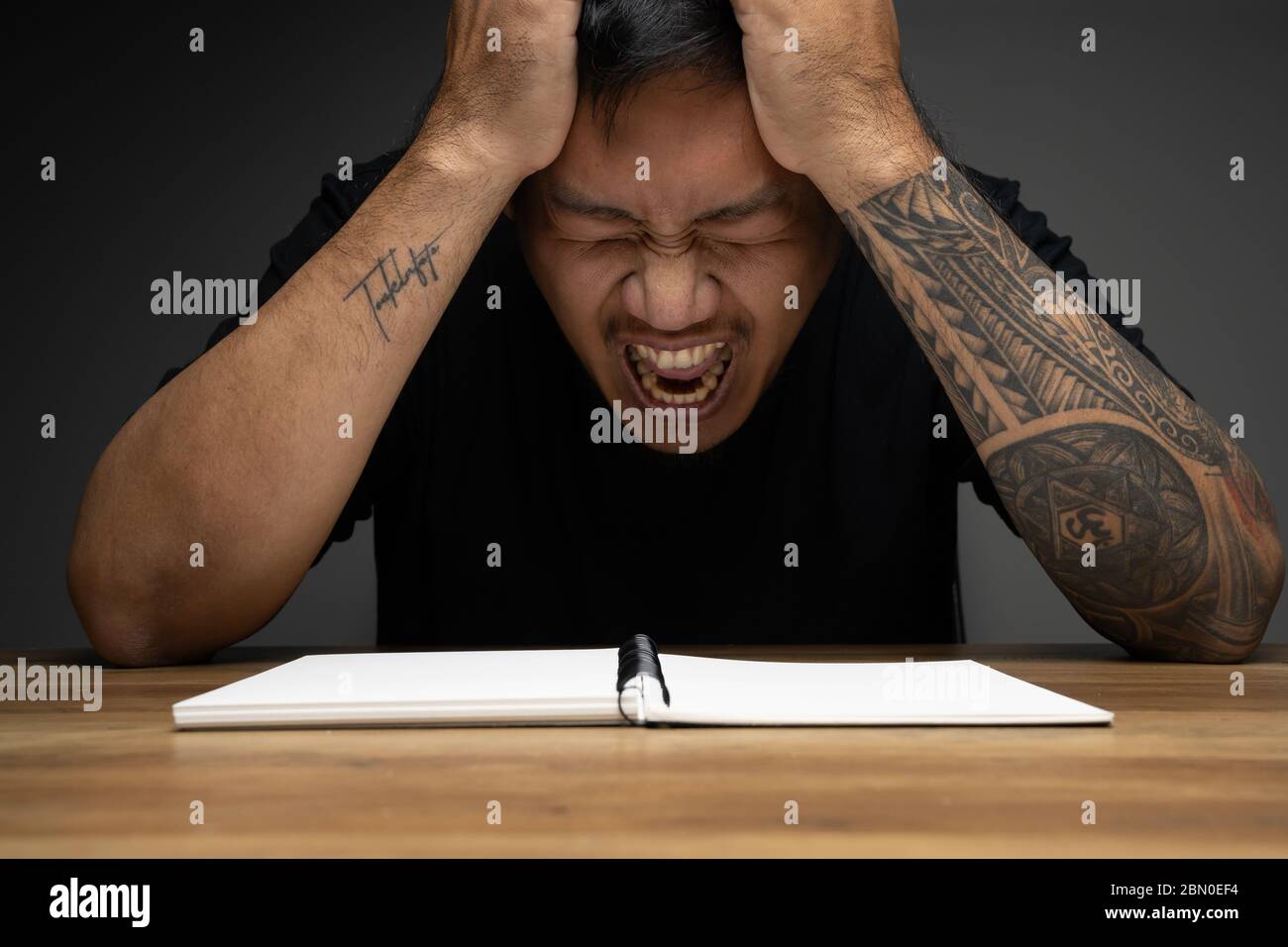 Asian Mann Schreiben auf einem Notizbuch mit wütenden Gesicht auf Holztisch. Konzept über Remote-Arbeit. Stockfoto