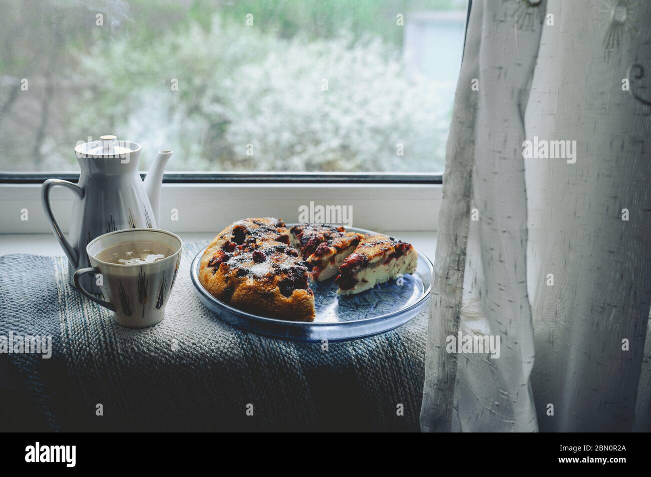Gemütliches Zuhause Tee-Party am Fenster bei schlechtem Wetter. Porzellan weiße Teekanne und Tasse mit Tee, hausgemachte Beerenkuchen auf einem runden blauen Teller gegen eine blühende tr Stockfoto