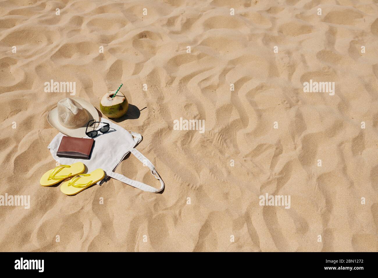 Leinenbeutel mit Flip-Flops, Strohhut, Sonnenbrille und grüner Kokosnuss mit Trinkhalm am Sandstrand Stockfoto