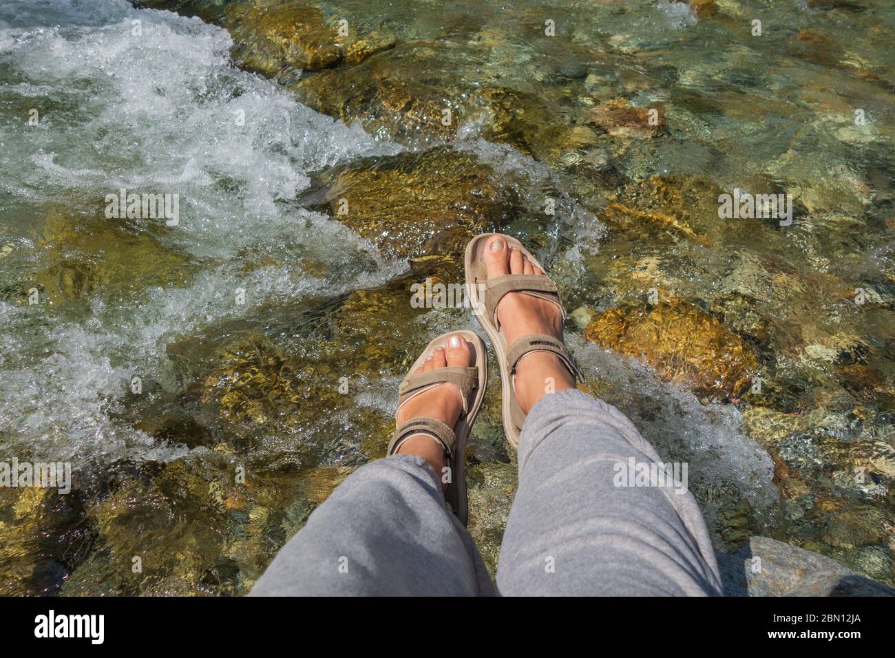 Gebräunte Beine der Frauen in offenen Sport-Sandalen auf einer Wanderung auf einem Bergfluss. Reisekonzept, das Konzept des Wanderens Stockfoto