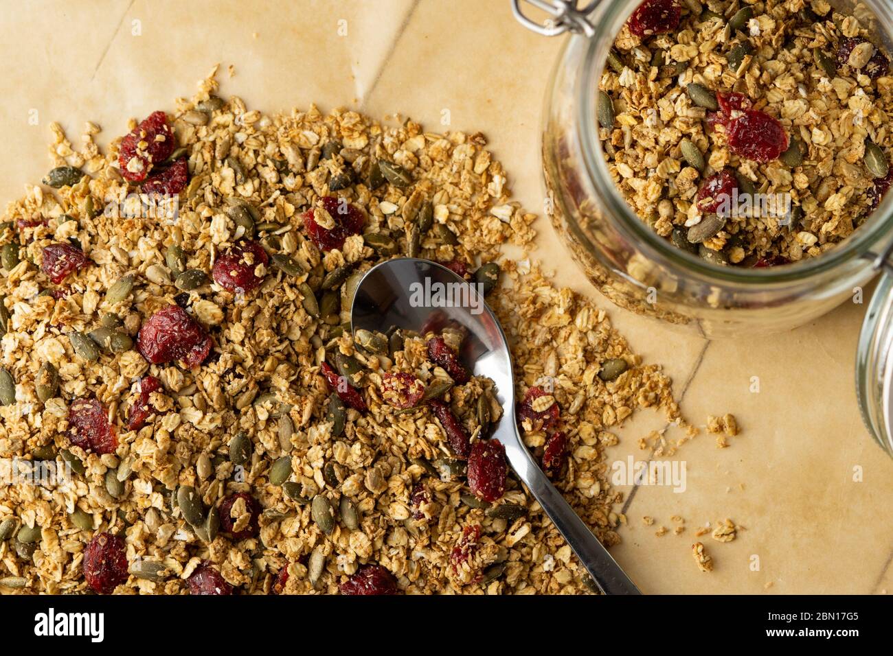 Hausgemachtes Müsli mit Samen und trockenen Cranberries auf einem Backpapier. Gesundes veganes Frühstück, Snack oder Mittagessen. Feiling Glas mit Metalllöffel. Draufsicht. Stockfoto