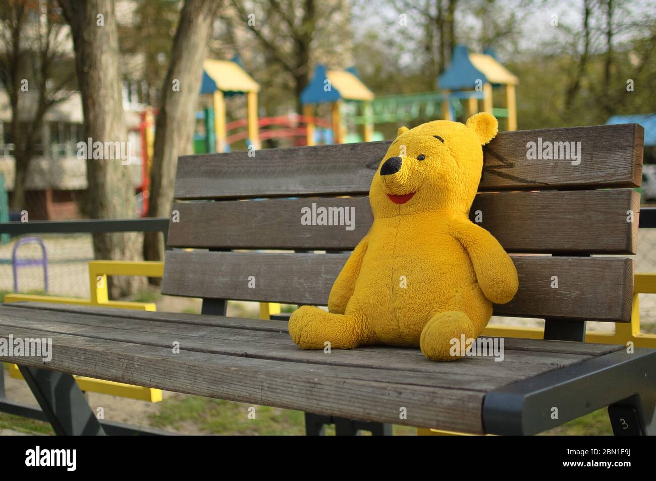 2020.04.18 Lviv, Ukraine. Gelbbär Spielzeug sitzt auf der Bank in der Nähe von leeren geschlossenen Spielplatz im Freien ohne Kinder wegen Quarantäne Maßnahmen Stockfoto