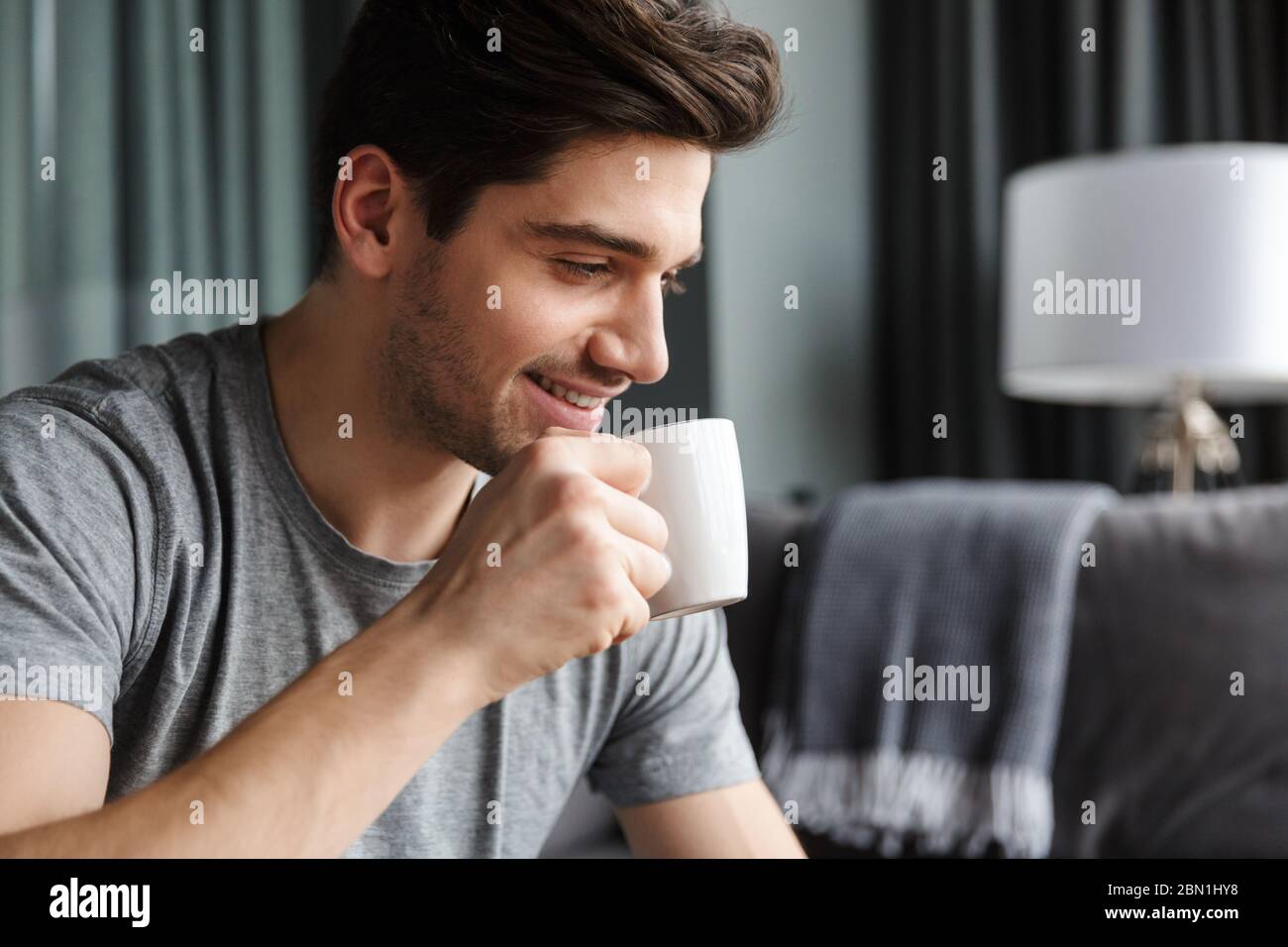 Nahaufnahme eines lächelnden jungen bärtigen Mannes, der Tee aus einer Tasse im Wohnzimmer trinkt Stockfoto
