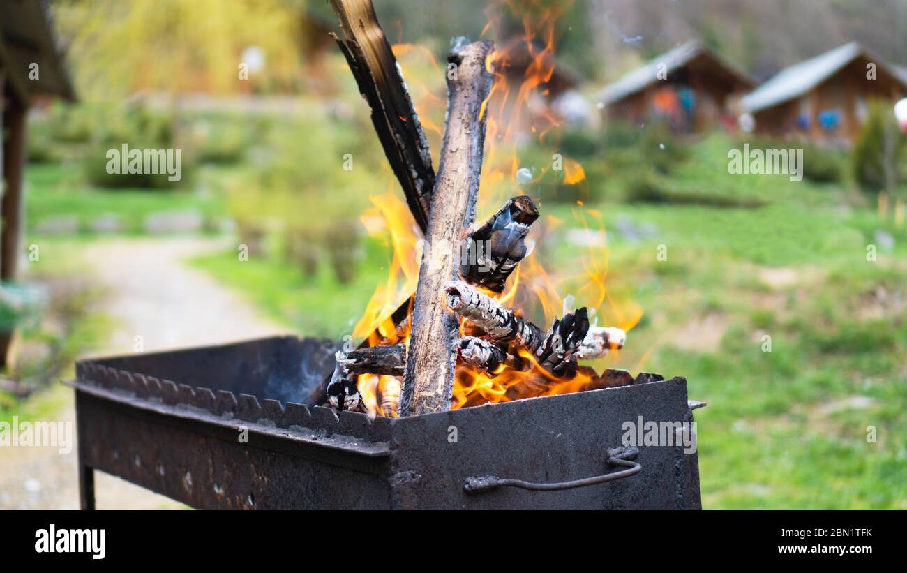 Metall schwarz Grill und brennendes Holz innen und Feuer auf grünem Hintergrund in der Nähe des Hauses. Bereiten Sie sich auf das Grillen vor Stockfoto