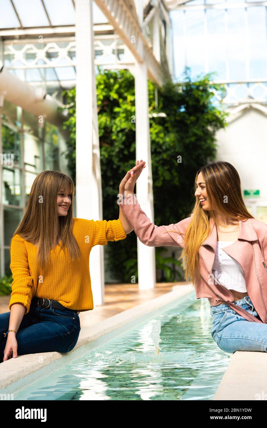 Zwei attraktive junge kaukasische Mädchen mit blonden Haaren schütteln die Hände mit ihren Armen in der Luft lächelnd an einem Brunnen in einem hell erleuchteten Raum mit dem Stockfoto