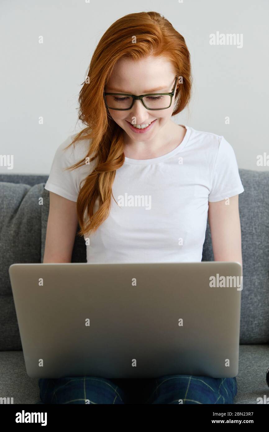 Rotschopf Mädchen sitzt auf einem Sofa mit weißem T-Shirt und Brille, lächelnd, tippen auf einem Laptop. Stockfoto