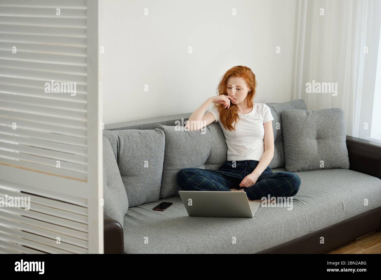 Ein rothaariges Mädchen in Heimkleidung sitzt auf einem Sofa und sieht nachdenklich in einen Laptop vor ihr. Stockfoto