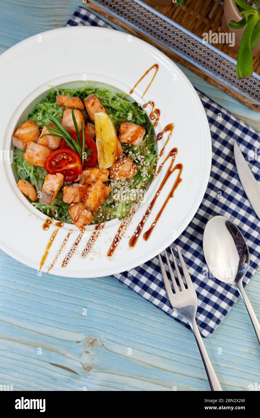 Reisnudeln mit Lachs und Spinat. Mittagessen in einem weißen Teller auf dem Tisch. Tomaten, Zitrone, Fisch, Rosmarin, Soi und Sesam. Stillleben im Essen Stockfoto