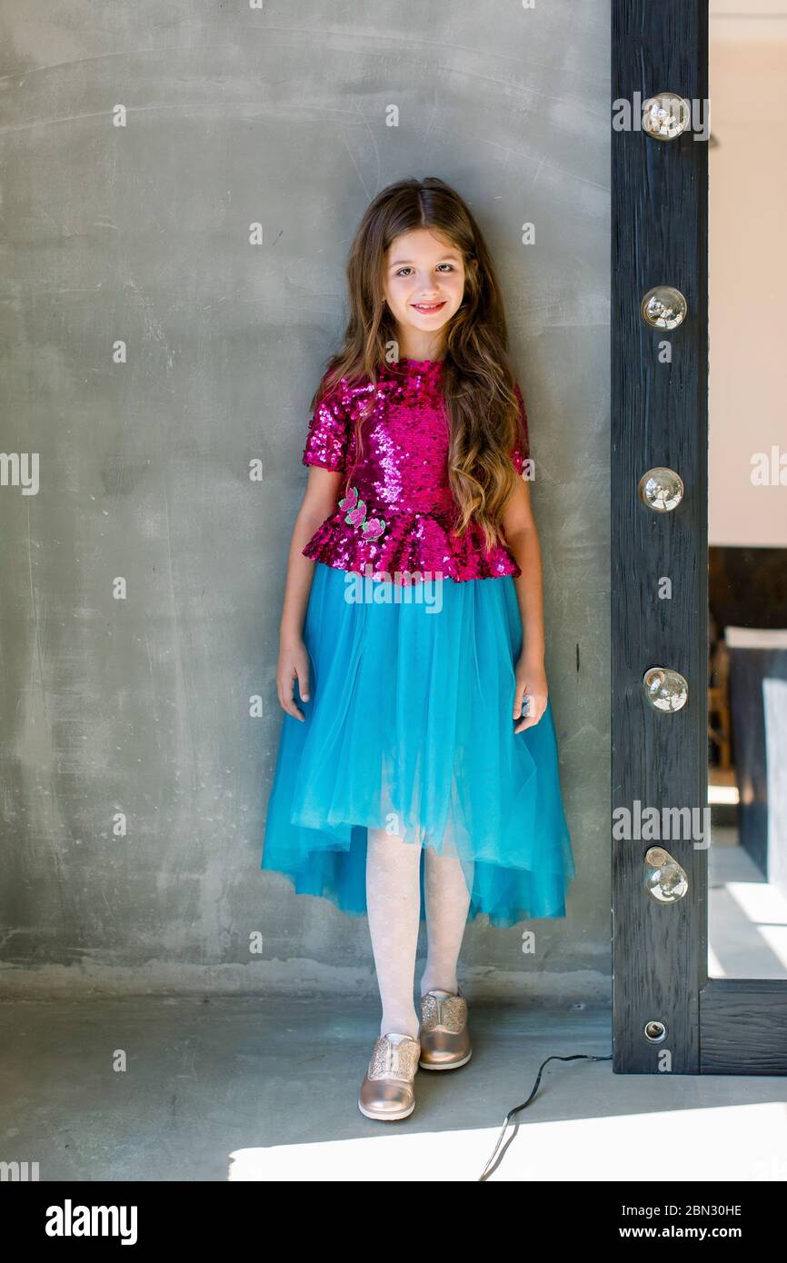 Schöne niedliche kleine Mädchen in einer rosa glänzenden Bluse und einem blauen Rock auf einem grauen Hintergrund stehen in der Nähe Spiegel. Stockfoto