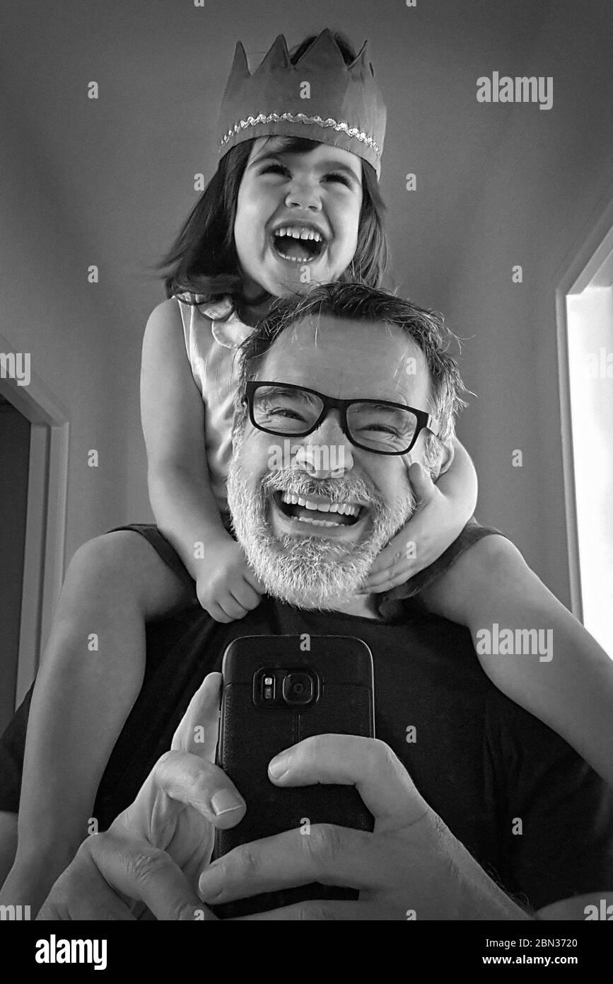 Selfie eines lächelnden Vaters, der seine Tochter auf den Schultern vor einem Spiegel trägt. Stockfoto