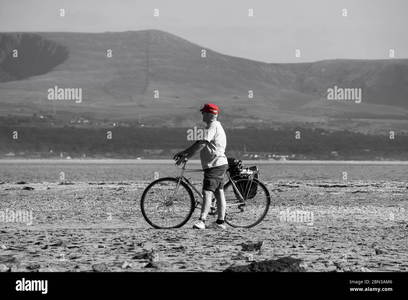 Mann in roter Mütze, britischer Urlauber, isoliert am leeren britischen Strand, der sein Fahrrad auf Sand schiebt. Monochromer Hintergrund. Urlaub im Sommer. Stockfoto