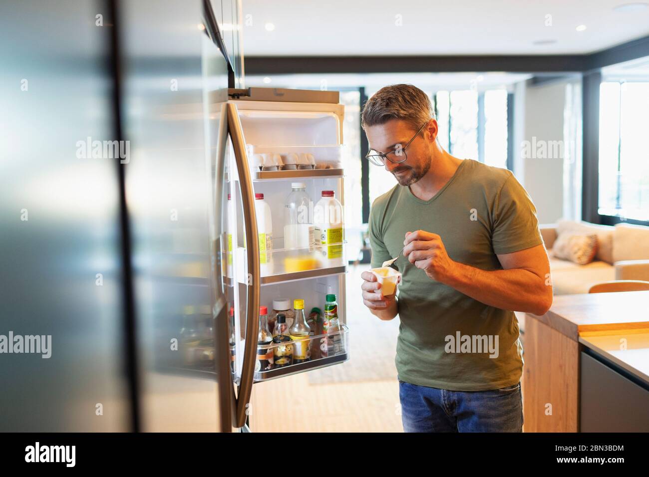 Mann, der Joghurt im offenen Kühlschrank in der Küche isst Stockfoto