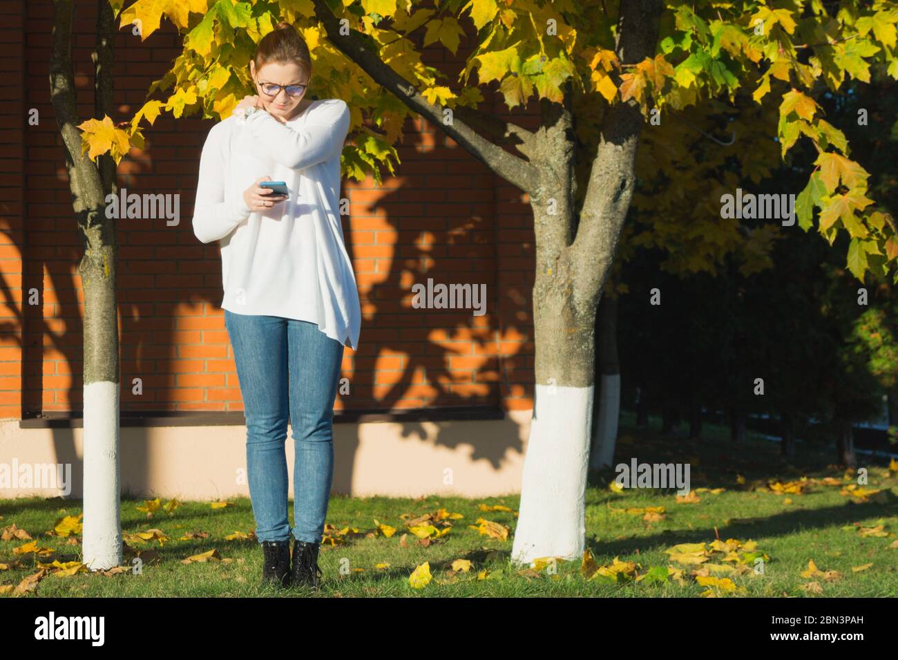 Eine junge Frau runzelt ihr Smartphone beim SMS-Schreiben mit der Stirn. Sie reibt sich ihre Schulter in der Hoffnung, ihre Nackenschmerzen zu lindern. Smartphone-Einbruch. Stockfoto