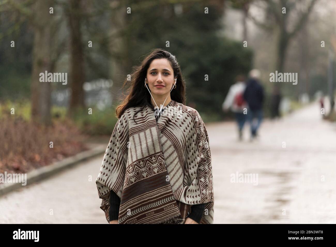 Junge Frau posiert auf Parkstraße gekleidet in Poncho. Vorderansicht. Wegschauen. Stockfoto