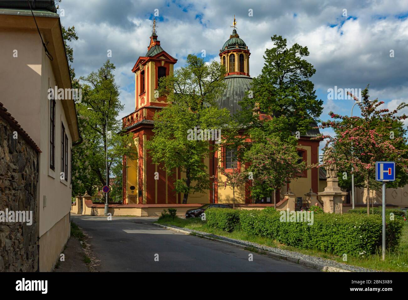 Kunratice, Prag / Tschechische Republik - 6. Mai 2020: Die barocke Kirche des Hl. Jakobus des Großen mit rot-gelber Fassade, umgeben von grünen Bäumen. Stockfoto