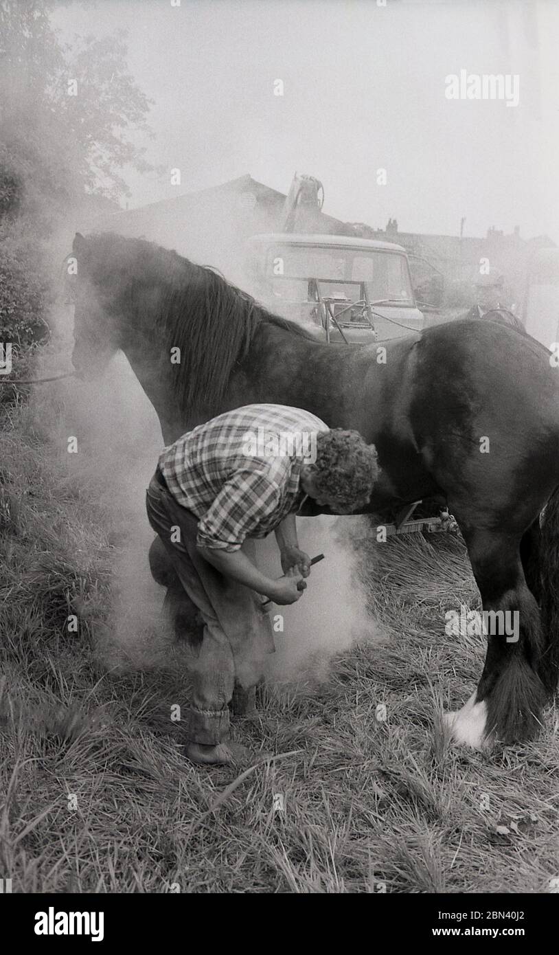 1987, heißes Shoeing..... Auf einer Zigeuner- oder Reisesternmesse steigt Dampf auf, während ein Farrier ein Pferd umhackt und es auf den Haufen legt, während es noch rot heiß ist, damit es an einem Ort brennt, an dem es richtig passt, England, Großbritannien. Stockfoto