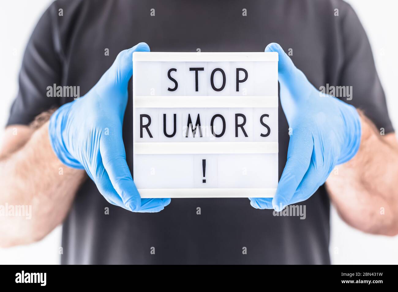 Fake News Infosemics during Covid-19 Pandemie Concept. Mann trägt Schutzmaske und medizinische Handschuhe an den Händen halten Leuchtkasten mit Text Stop Rumors. Stockfoto