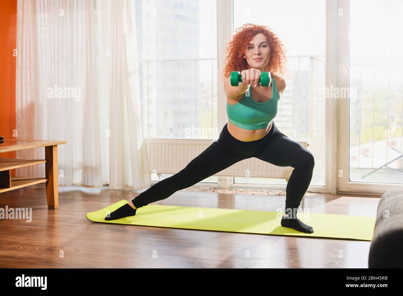Junge positive Frau halten Hanteln und Training, machen Ausfallschritte Übungen zu Hause Innenraum während der Welt covid Isolation Stockfoto