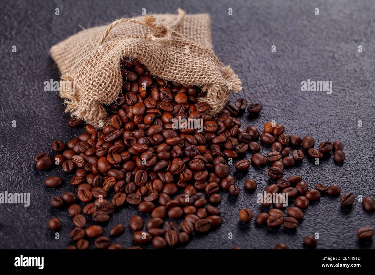 Geröstete, aromatische Kaffeebohnen, die aus einem Leinenbeutel auf einer dunklen Oberfläche bestreut wurden. Das Konzept der Tradition des Morgenkaffees. Ansicht von oben Stockfoto