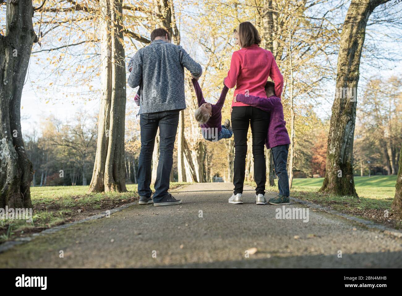 Blick von hinten auf eine junge Familie mit drei Kindern, die die Hände halten, während sie im Park spazieren, Mutter und Vater, die das Kind in der Mitte heben. Stockfoto