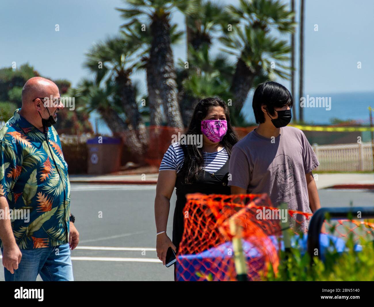 Ein junges Paar, das richtig Schutzmaske trägt, und ein Mann mittleren Alters, der seine Maske falsch unter seinem Kinn trägt, gehen in der Nähe des Ozeans. Stockfoto