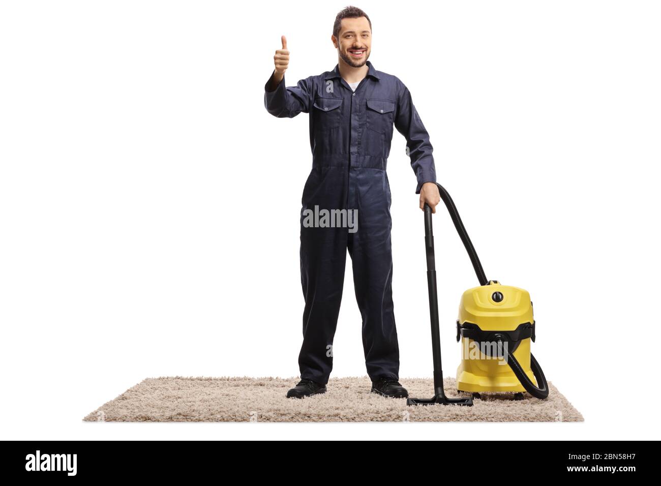 Ganzkörperportrait eines Hausmeisters in Uniform mit einem Staubsauger auf einem Teppich stehend mit Daumen nach oben isoliert auf weißem Hintergrund Stockfoto