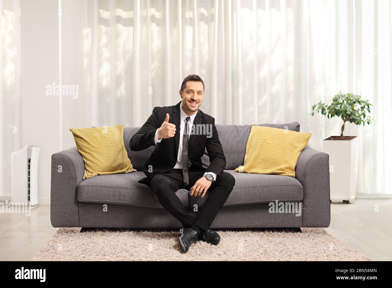 In voller Länge Porträt eines fröhlichen jungen Geschäftsmann sitzt auf einem Sofa und zeigt Daumen nach oben zu Hause Stockfoto