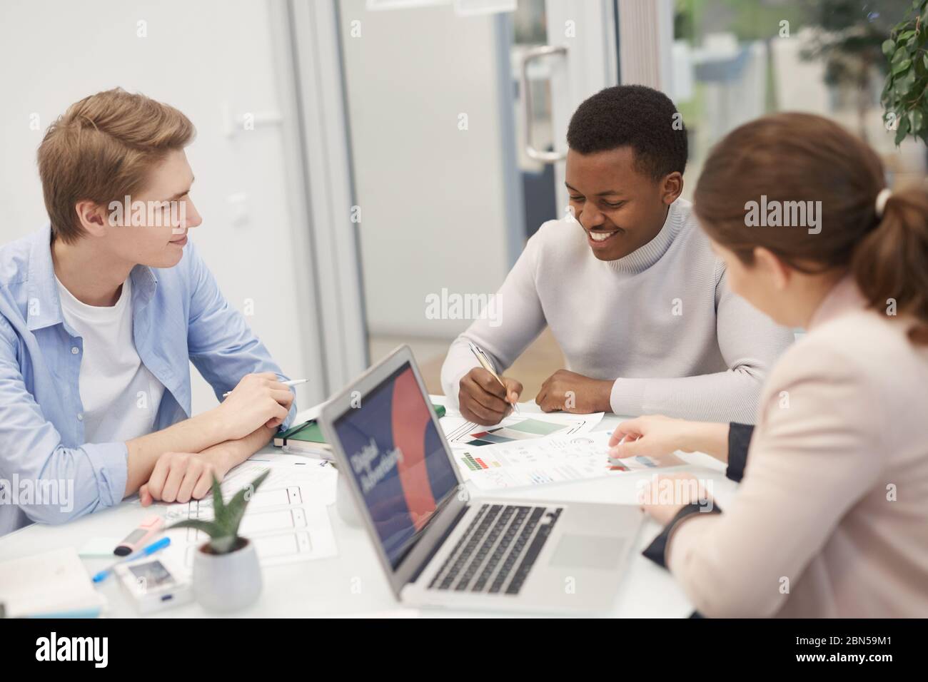 Gruppe junger Geschäftsleute, die über ein Arbeitsprojekt sprechen und fröhlich lächeln, während eines Treffens im Büro, Copy-Space Stockfoto