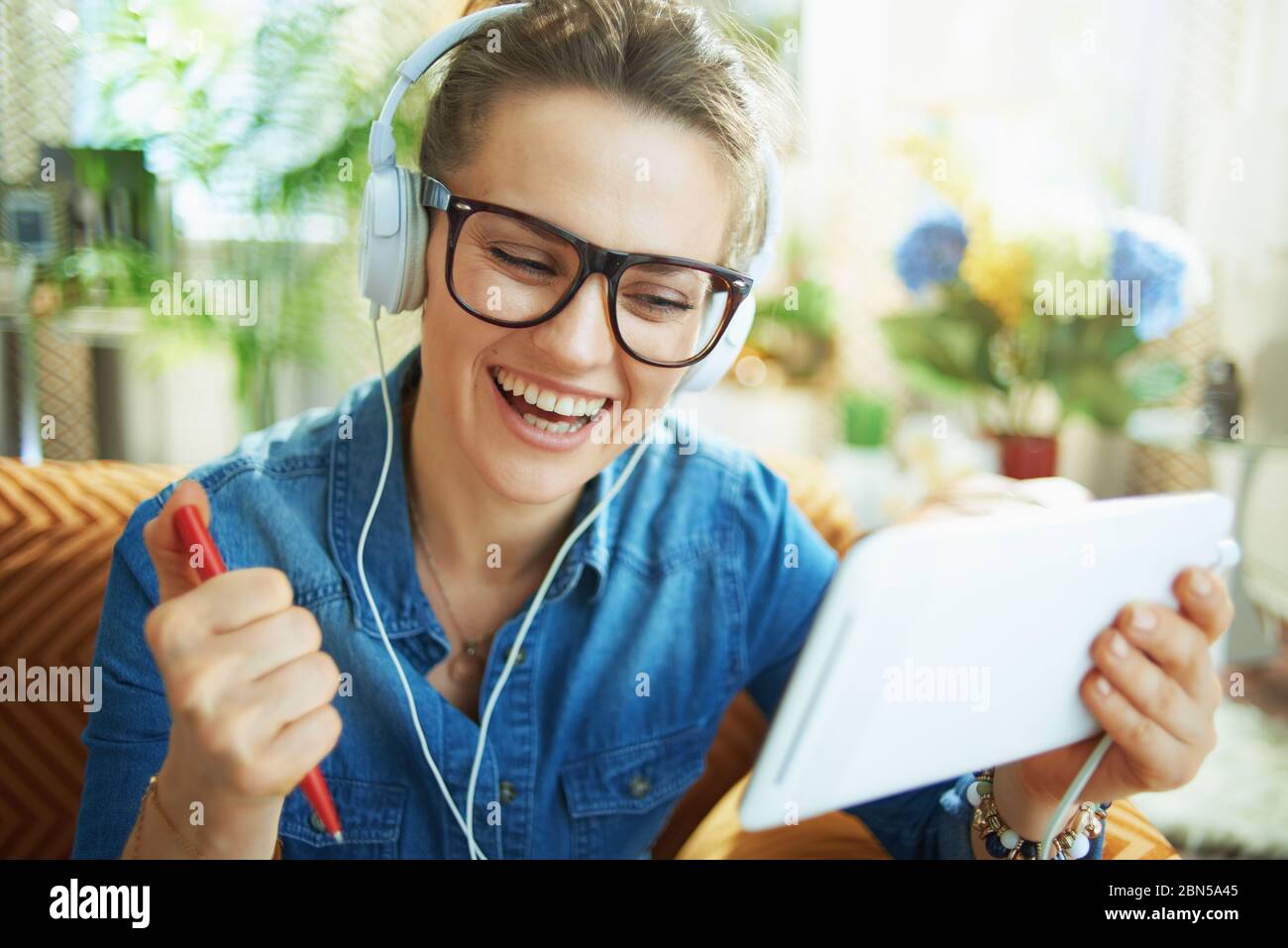 Happy stilvolle Frau in Jeans-Shirt mit weißen Kopfhörern und Tablet-PC studieren online in modernen Zuhause an sonnigen Tag. Stockfoto