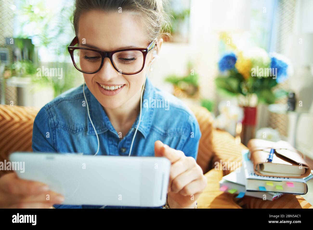 Lächelnde 40 Jahre alte Hausfrau im Jeans-Hemd mit weißen Kopfhörern und Tablet-PC studieren online im modernen Haus an sonnigen Tagen. Stockfoto