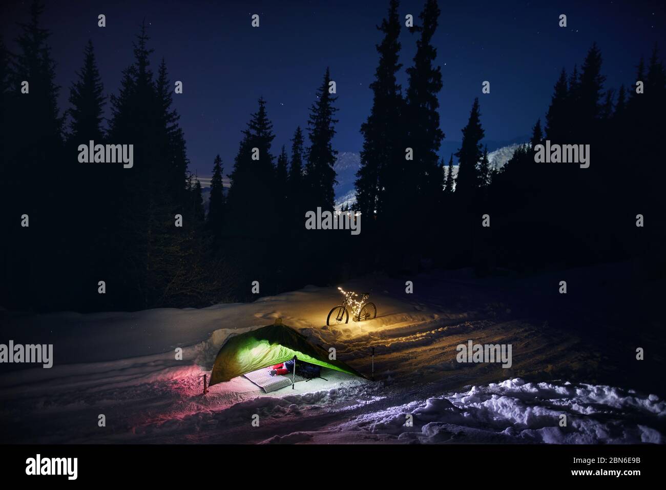 Leuchtende grüne Zelt und Fahrrad mit Weihnachtsbeleuchtung im Winter Wald unter Nachthimmel mit Sternen. Bikepacking Lebensstil und Weihnachten Konzept. Stockfoto
