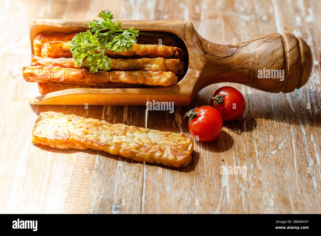 Gebratenes Tempeh in einer Holzkelle, mit Petersilie und Kirschtomaten verziert, auf Holzfläche. Stockfoto