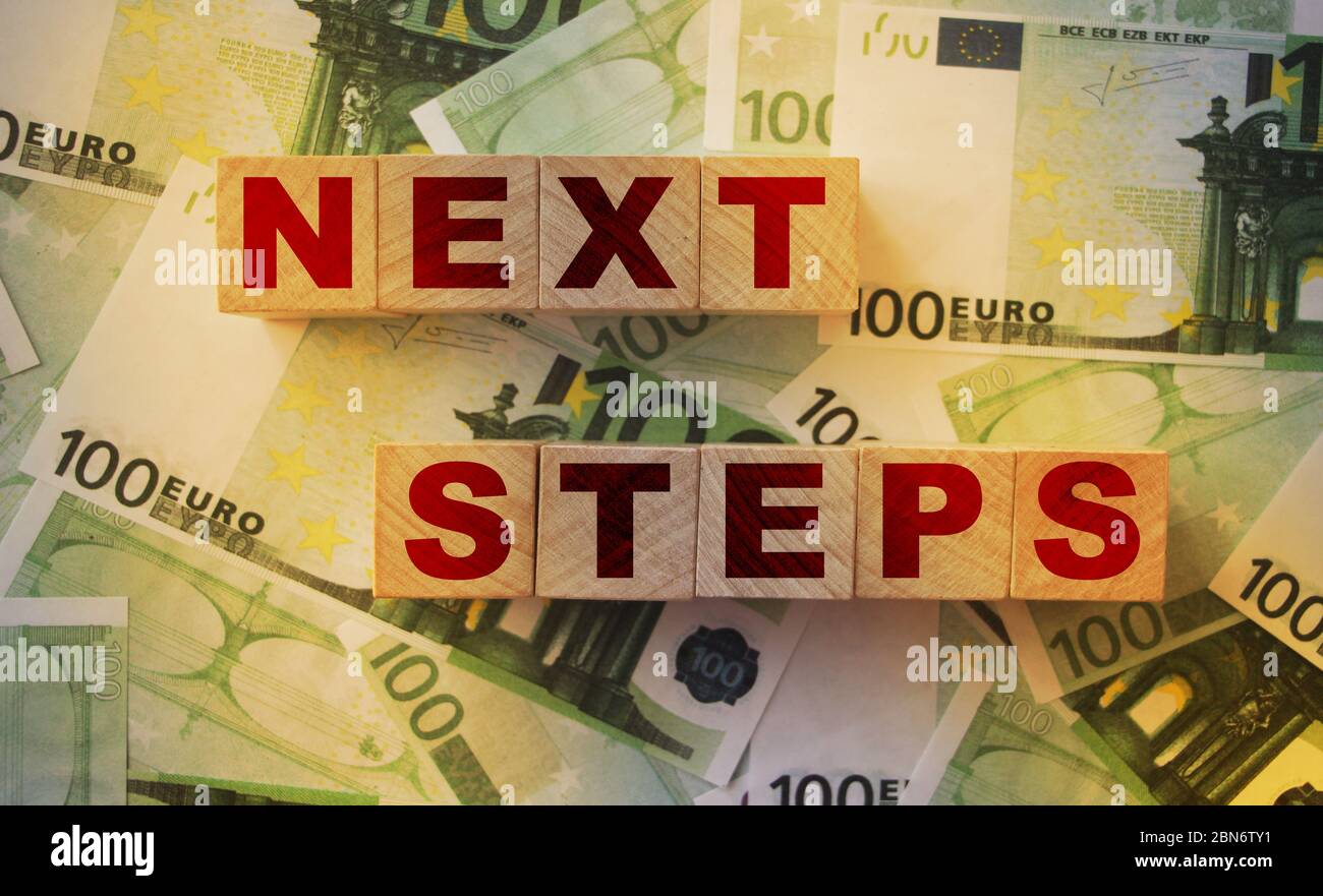 Die Worte Nächster Schritt auf kleinen Holzdices auf einem Tisch, voll von 100 Euro-Banknoten . Konzept der Geschäftsplanung Stockfoto