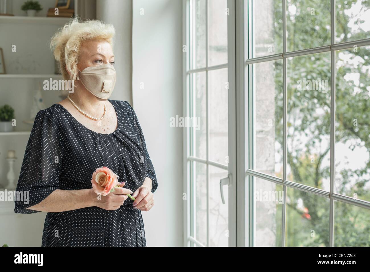 Ältere Frau mit Schutzmaske und schwarzem Kleid, die zu Hause auf das Fenster schaut Stockfoto