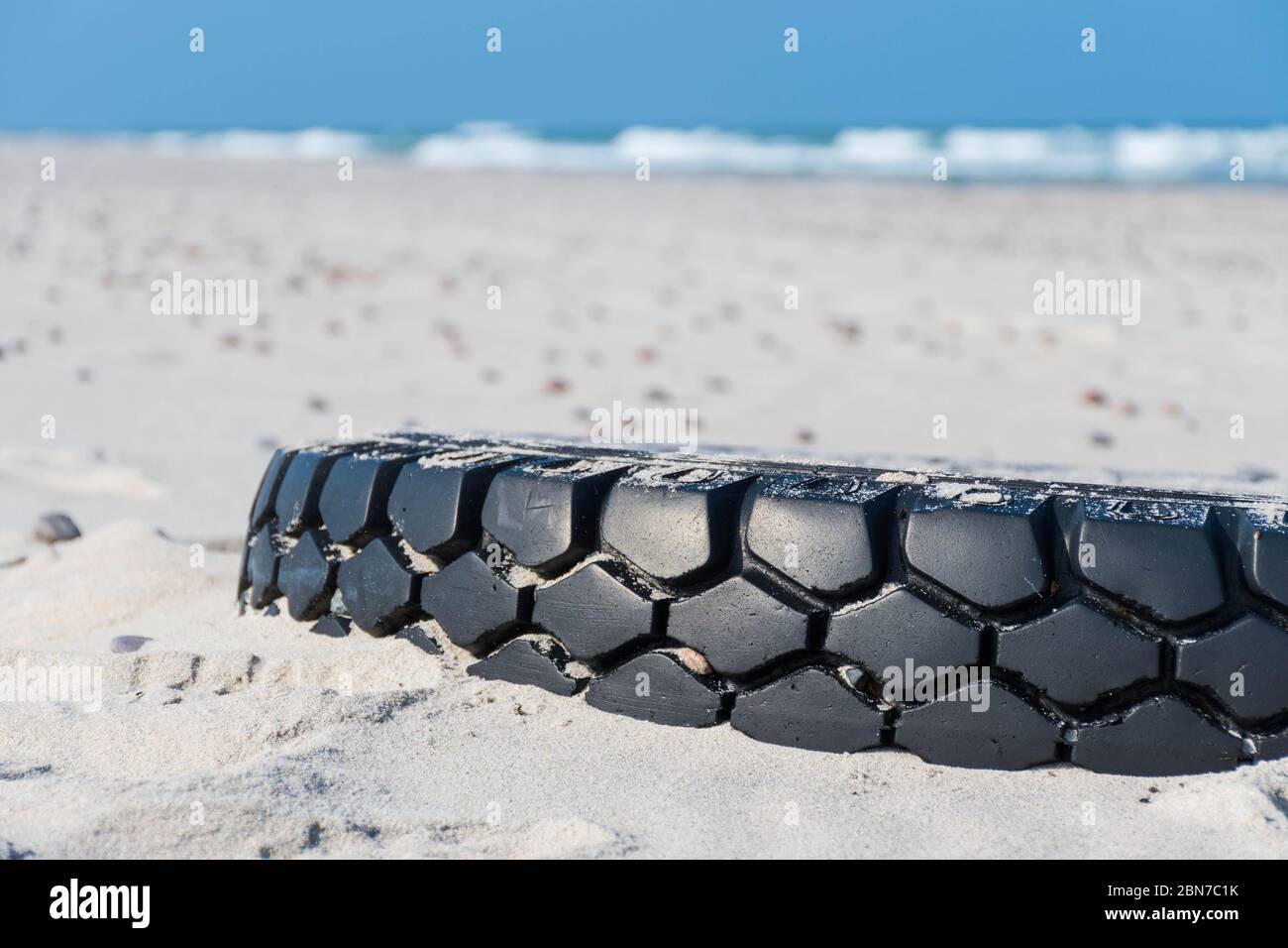 Großer schwarzer Gummireifen links auf einem Sandstrand mit blauem Meer und Wellen auf dem Hintergrund, Umweltverschmutzung Konzept Stockfoto