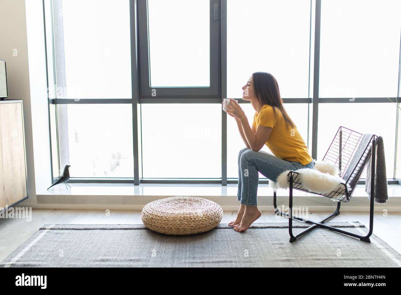 Junge Frau auf einem modernen Stuhl vor dem Fenster, die sich in ihrem Wohnzimmer entspannt und Kaffee oder Tee trinkt Stockfoto