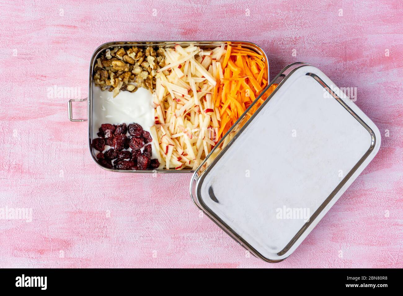Vegetarische Karotten und Apfelsalat in einem großen Edelstahl-Lebensmittelbehälter. Kunststoff-Lunchbox auf rosa Hintergrund isoliert. Keine Verschwendung, umweltfreundlich. Stockfoto