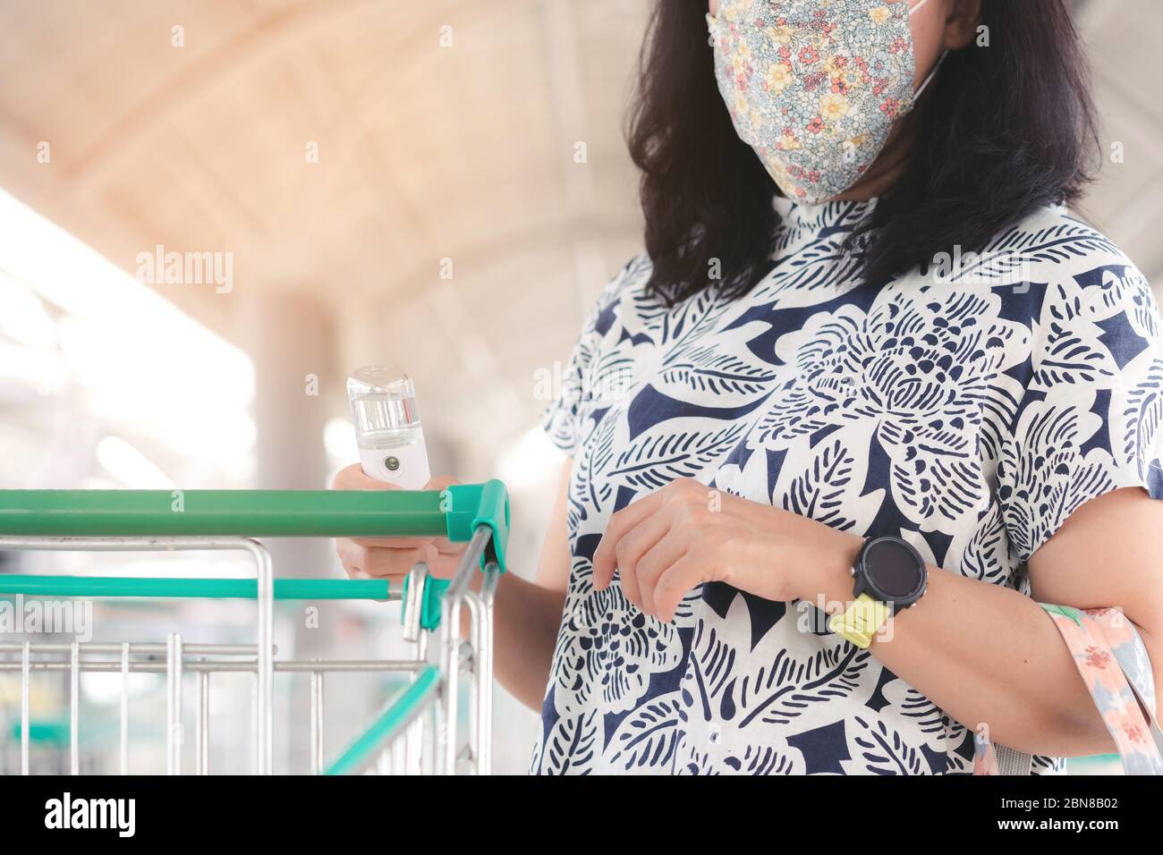 Frau tragen Maske mit Alkohol Nano-Sprühnebel antiseptische Reinigung auf Warenkorb Wagen Griff Schutz während Coronavirus Pandemie Covid-19. W Stockfoto