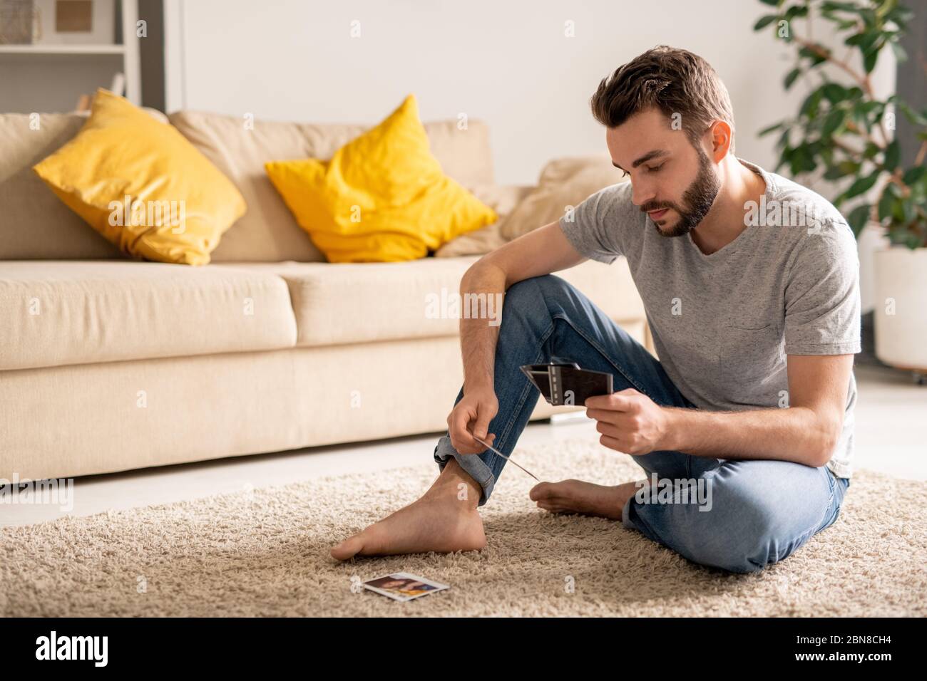 Trauriger junger Mann, der im Wohnzimmer auf einem Teppich sitzt und Fotos durchschaut, während er sich an das Leben vor der Coronavirus-Epidemie erinnert Stockfoto