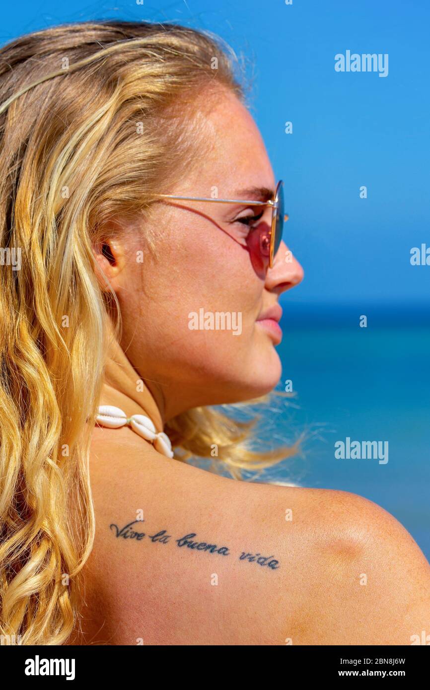 Porträt einer jungen blonden Frau mit Tattoo 'lebe das gute Leben' auf spanisch auf der Schulter Stockfoto